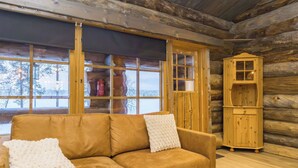 A log cabin interior with a tan leather sofa, a wooden cabinet, and large windows offering a view of trees. - Lumikenttien kutsu by Interhome (Kuusamo)