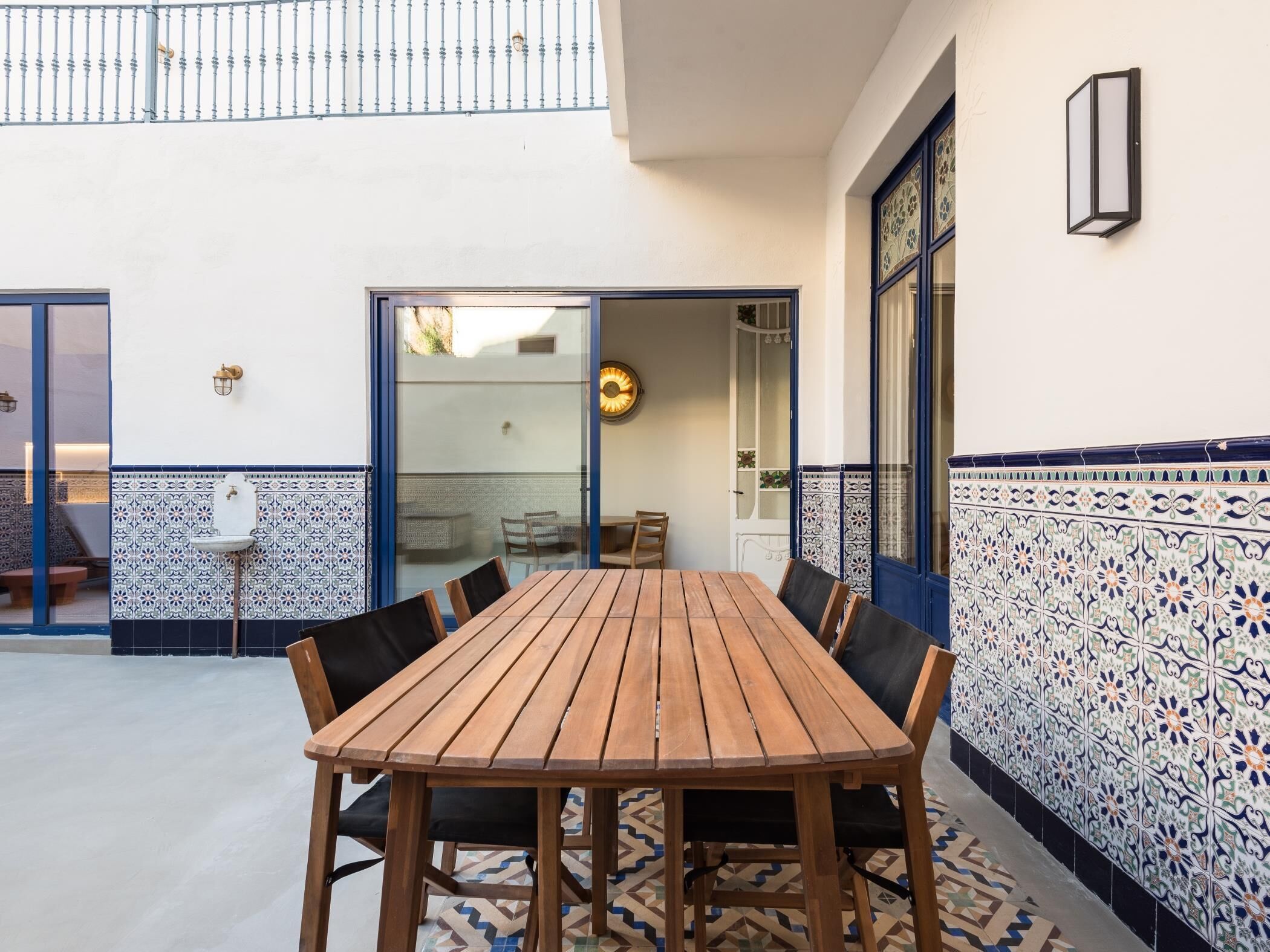 A wooden table with chairs in a courtyard area with patterned tiles and a glass door leading to an interior space.