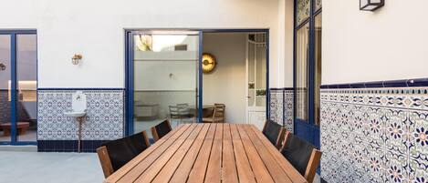 A wooden table with chairs in a courtyard area with patterned tiles and a glass door leading to an interior space.