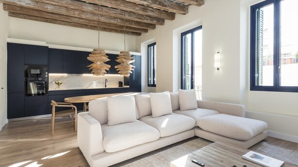 A modern living room with a sectional sofa, a wooden coffee table, and a kitchen area with dark cabinets and a hanging light fixture. - The Residences At Bartomeu 26, Suite Maricel, Spacious Elegance & Comfort (Sitges)