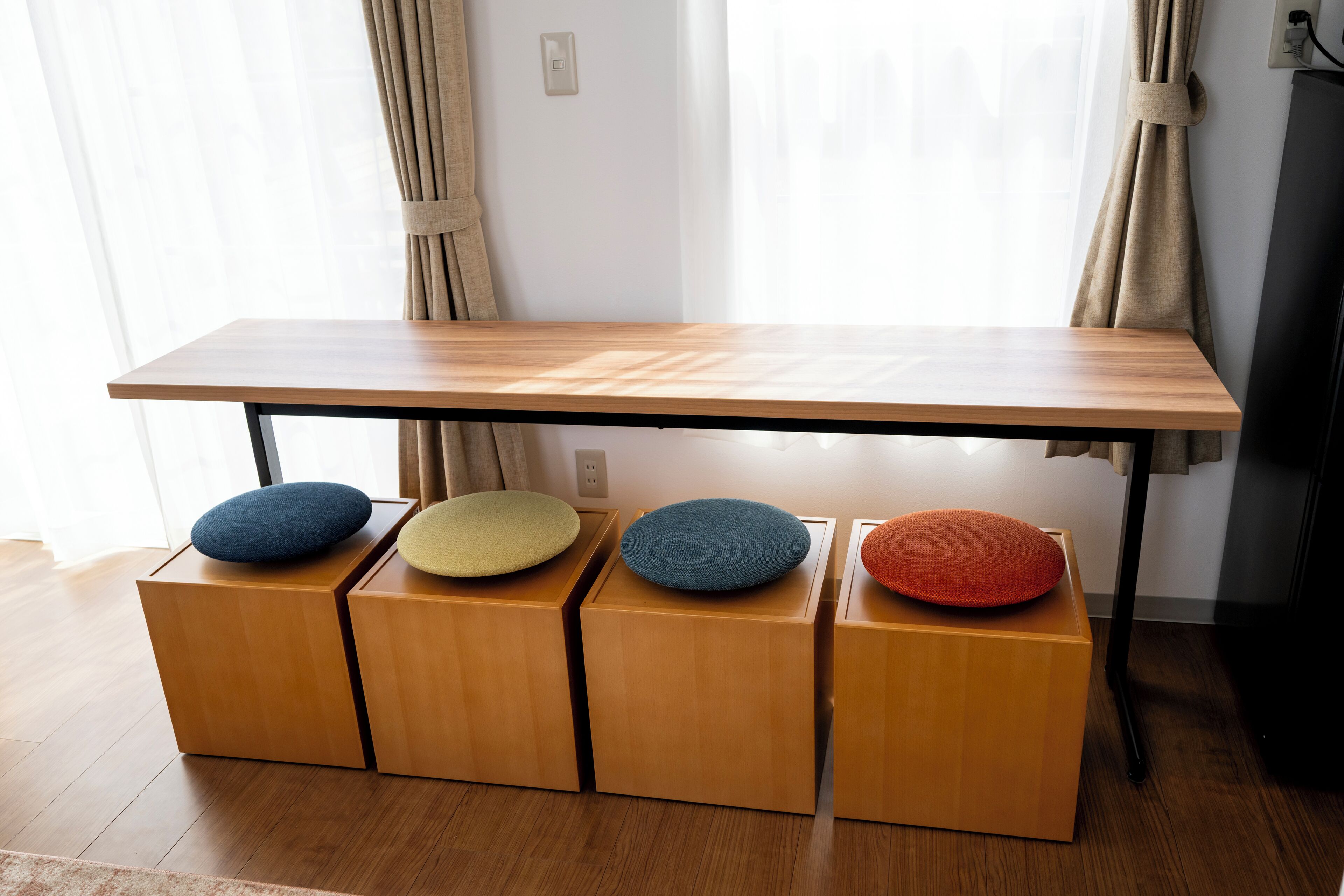 A wooden table with four matching ottomans, each with a different colored cushion, positioned in front of a window with sheer curtains.