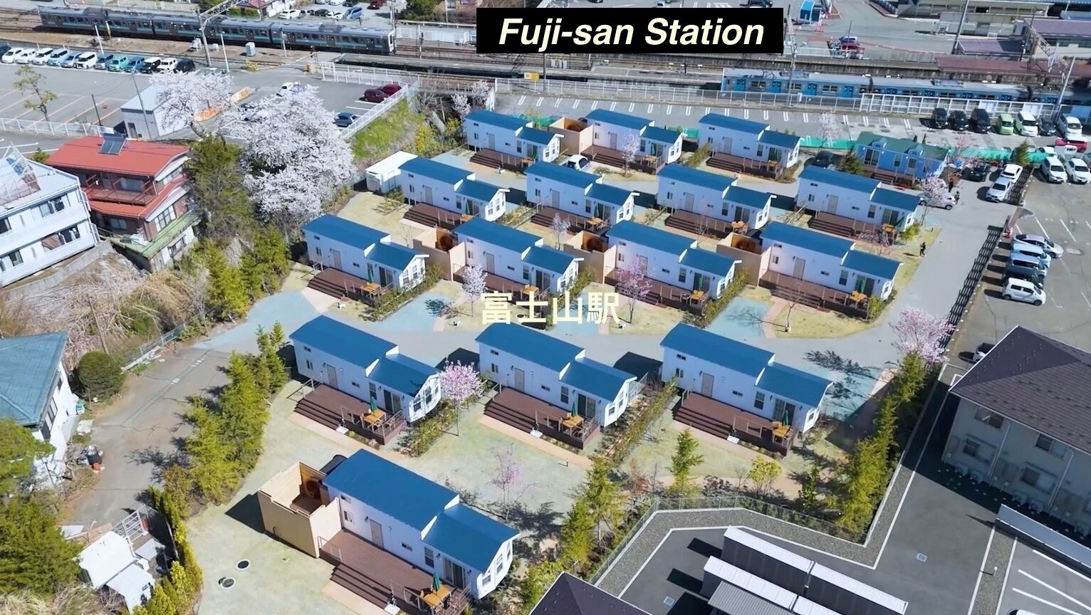 An aerial view of a residential area with multiple two-story houses, a train station in the background, and a parking lot.