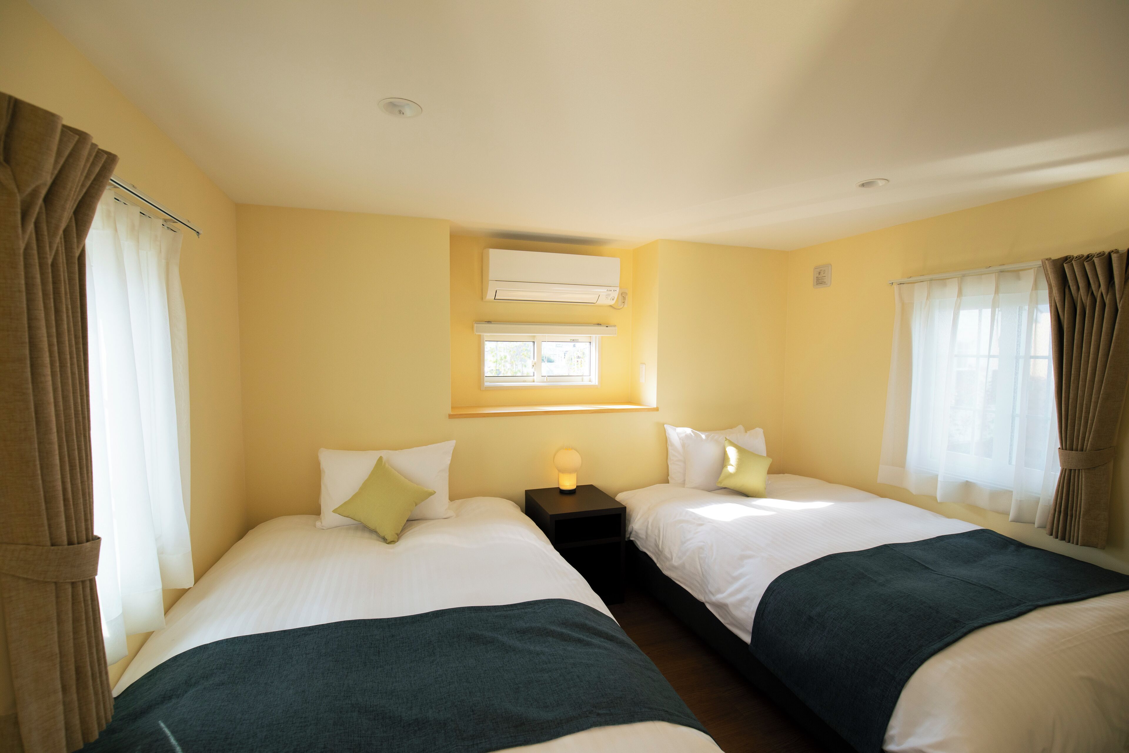 Camera d'albergo con due letti, una finestra con tende, un condizionatore e un comodino con lampada.