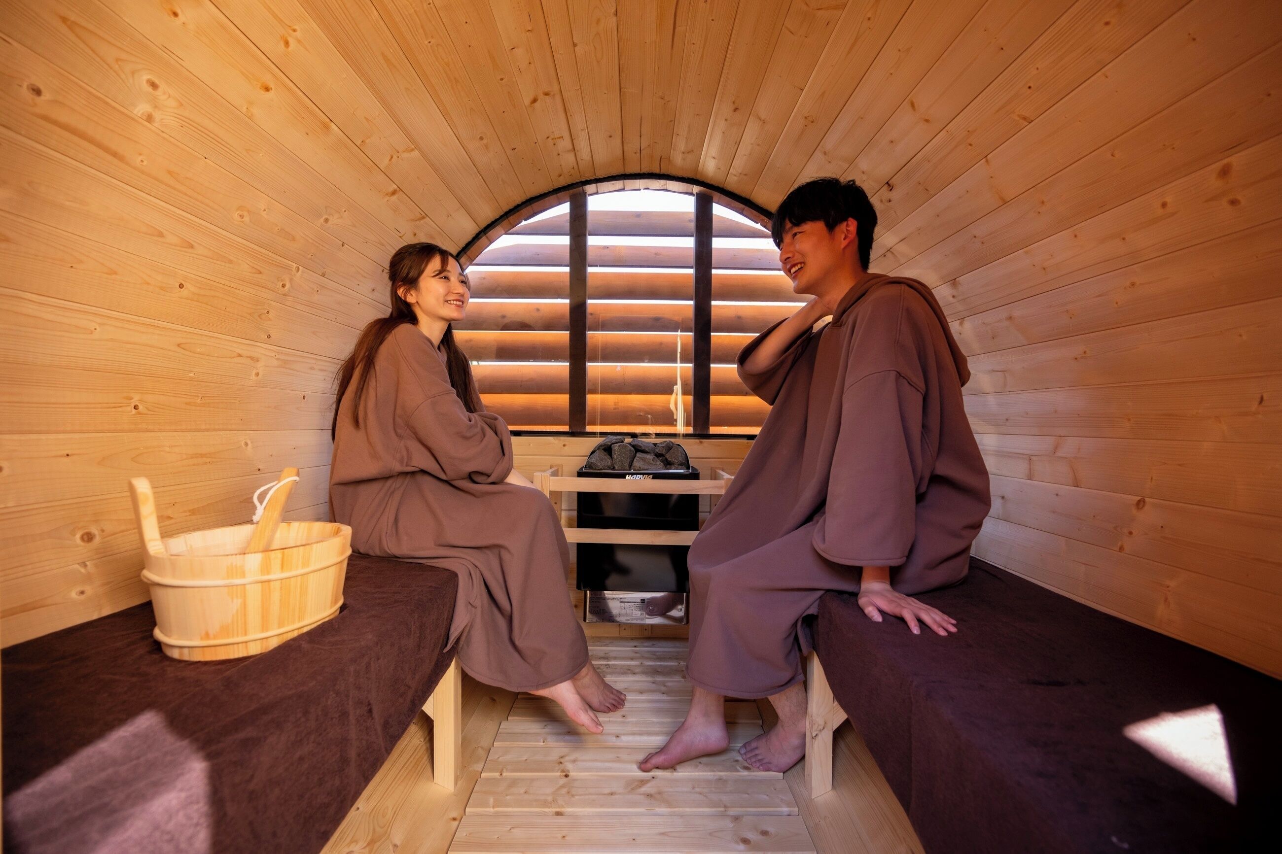 A sauna room with wooden walls and benches, a wooden bucket, and a shelf with towels.