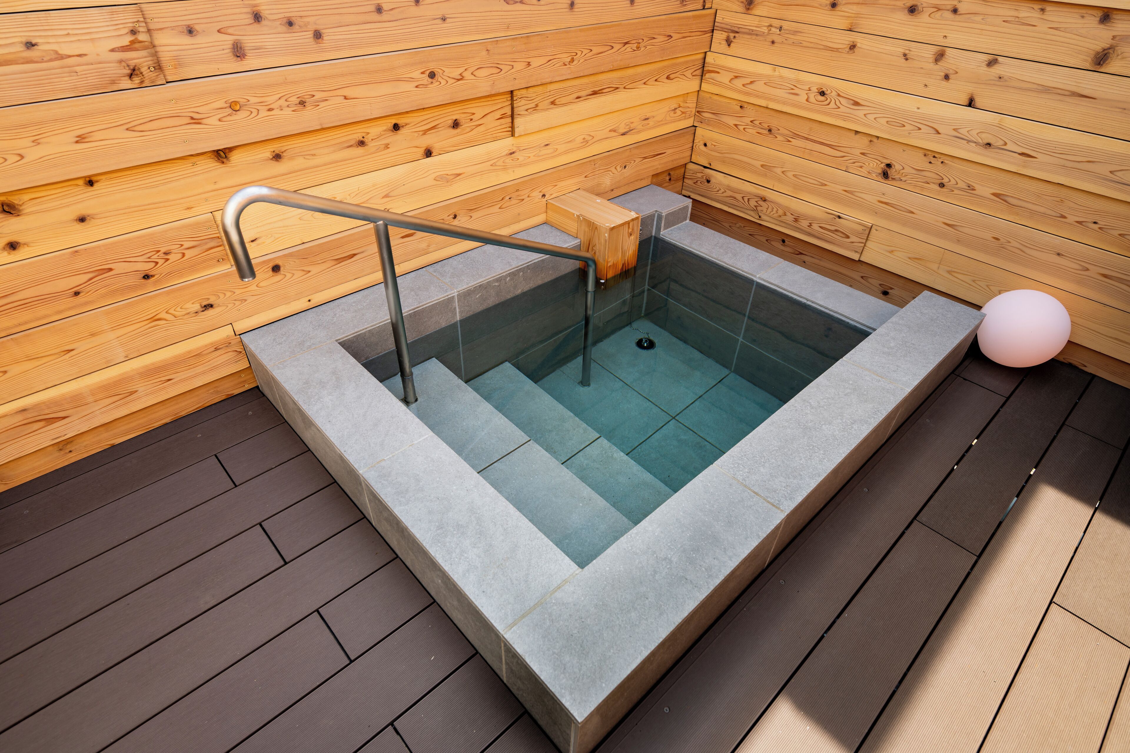 A wooden sauna with a glass-enclosed hot tub, a metal handrail, and a pink spherical light.