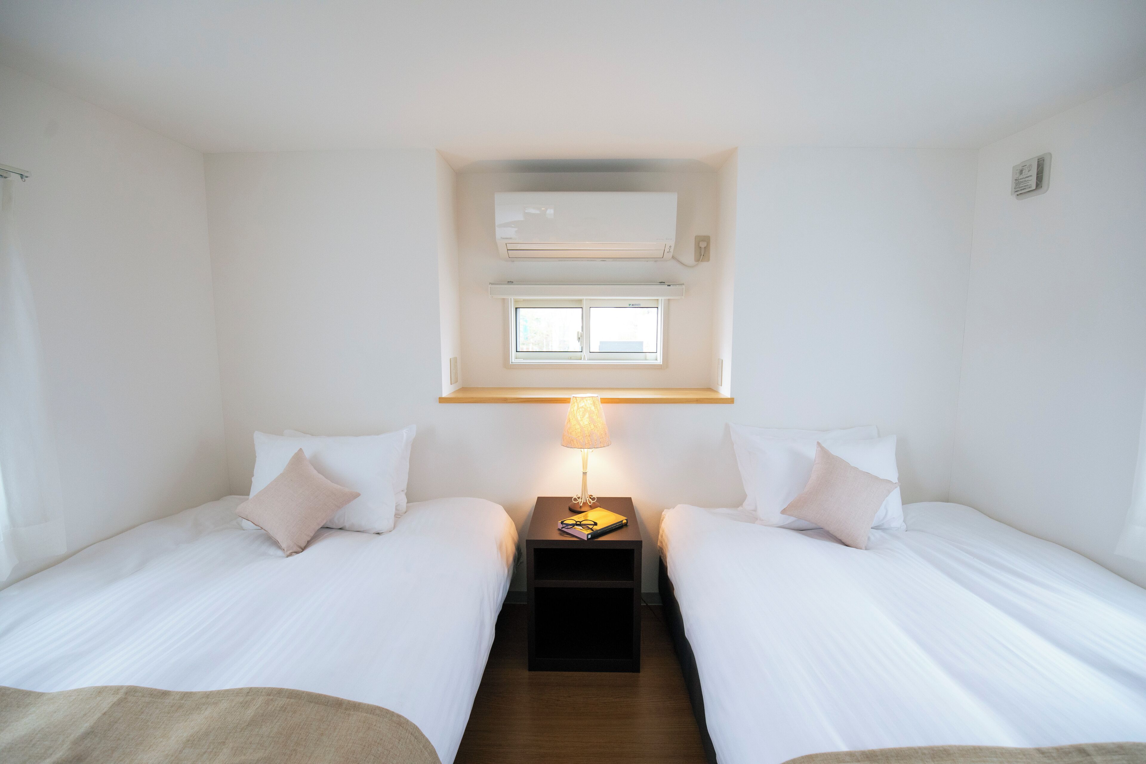 A hotel room with two single beds, a nightstand with a lamp, an air conditioning unit, and a window.