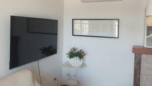 A living room with a flat-screen TV mounted on the wall, a small white side table with a vase of flowers, and a fireplace with a decorative piece. - Holidays in Mallorca: renovated apartment with sea views (Portocolom)