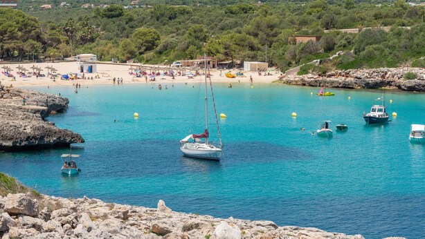 A coastal beach scene with clear turquoise waters, rocky shorelines, and a small sailboat anchored near the shore. - Holidays in Mallorca: renovated apartment with sea views (Portocolom)