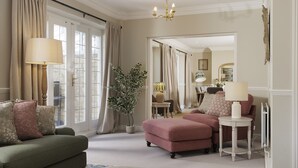 Ein gut beleuchtetes Wohnzimmer mit einem grünen Sofa, einem rosa Sessel und einem weißen Beistelltisch. Es gibt ein großes Fenster mit Vorhängen, eine Chandelüster und einen Spiegel an der Wand.
