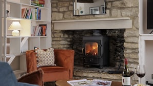 A cozy living room with a stone fireplace, a comfortable armchair, a coffee table with magazines, and a wine bottle. - Dog friendly cottage - The Roost (Coln St Aldwyns)