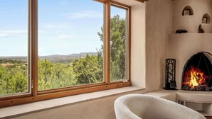 Una habitación con un ventanal que ofrece vistas a árboles y colinas. Hay una chimenea encendida y una silla blanca en la esquina.