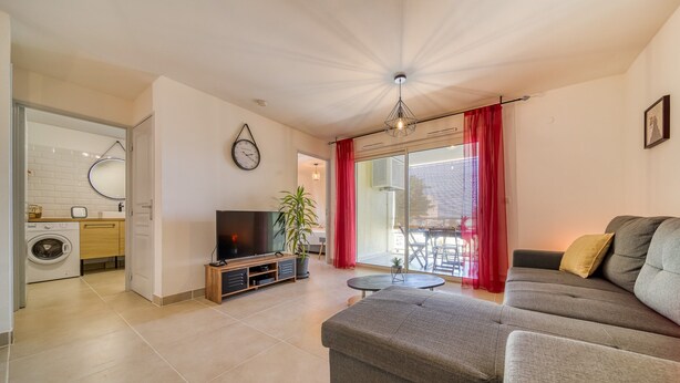 Una sala de estar moderna con un sofá gris, un televisor de pantalla plana, un mueble de madera pequeño, una planta en maceta y un suelo de vidrio corredera que da a un balcón.