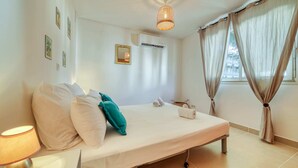 Un dormitorio ordenado con cama, almohadas, lámpara de noche, equipo de aire acondicionado y ventana con cortinas.