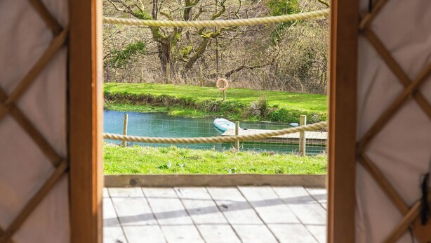 A view from inside a wooden-framed room with a rope barrier, overlooking a pond with a small boat and a lifebuoy. - Bracken Yurt - UKC6941 (Beaminster)