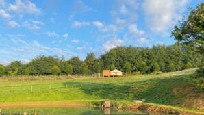A serene landscape with a small pond, a wooden cabin, and a forested hill in the background. - Bracken Yurt - UKC6941 (Beaminster)