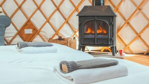 A cozy room with a wood-burning stove, a bed with neatly folded towels, and a wooden wall panel. - Bracken Yurt - UKC6941 (Beaminster)