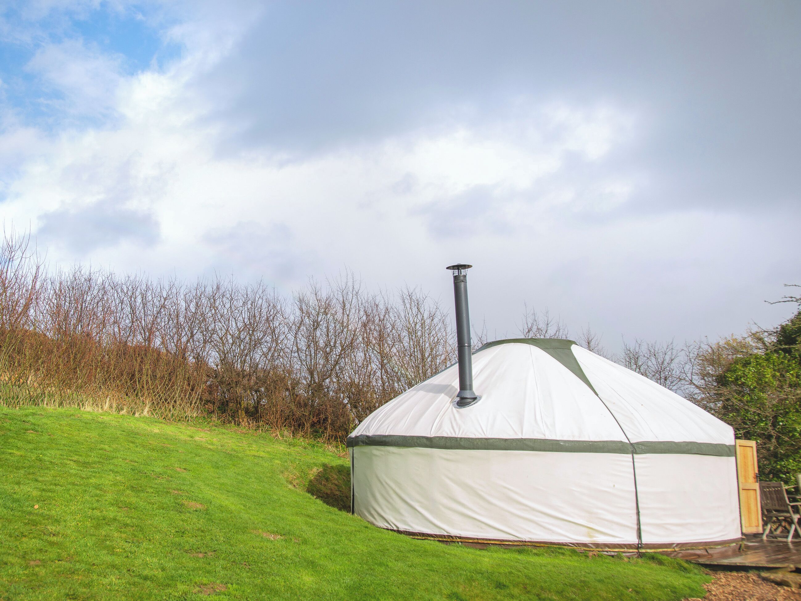 A white yurt with a chimney, set on a grassy hill with trees in the background.