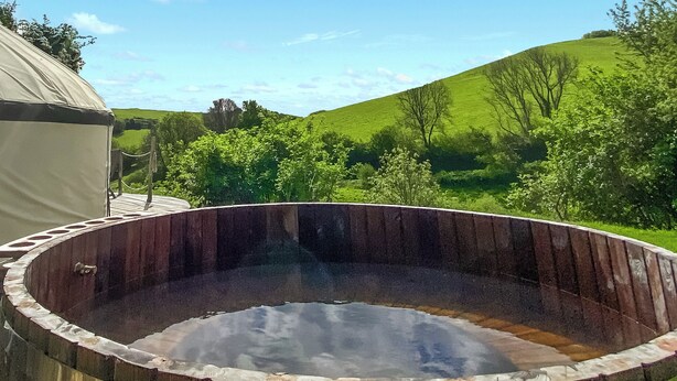 A hot tub with a wooden exterior, surrounded by a grassy landscape and trees. - Skylark Yurt - UKC6946 (Beaminster)