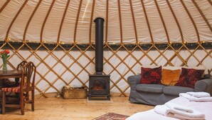 A cozy interior of a yurt with a central wood-burning stove, a sofa with patterned cushions, a wooden chair, and a dining table. - Skylark Yurt - UKC6946 (Beaminster)