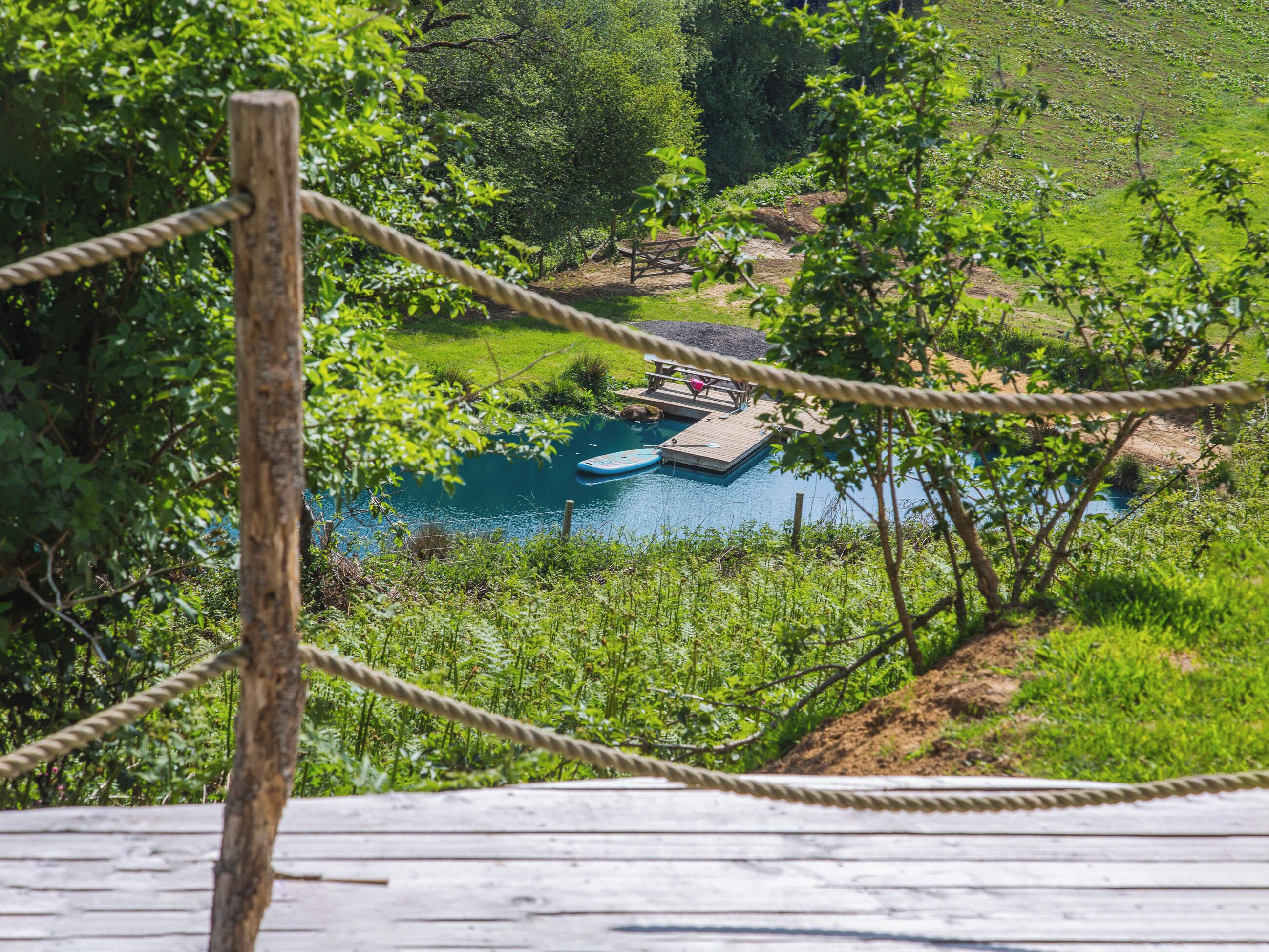 A wooden deck overlooking a clear blue pool with a wooden dock and lounge chairs, surrounded by lush greenery.