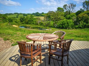 A wooden deck with a round table and four chairs, overlooking a lush green landscape with a pond and trees. - Hazel Roundhouse - UKC6942 (Beaminster)