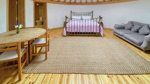 A room with a wooden ceiling, a bed, a gray sofa, a round wooden table, and a woven rug. - Mistle Roundhouse - UKC6943 (Beaminster)