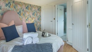 Une chambre à coucher avec un lit, des oreillers, une couverture, un mur orné d’un motif floral et une porte donnant sur une autre pièce.