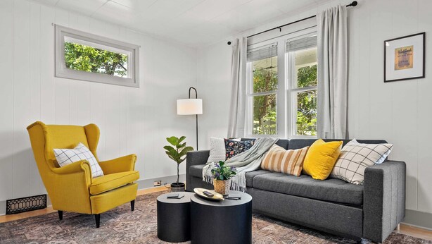 Un salon avec un fauteuil jaune, un canapé gris, deux tables d’appoint noires, une lampe sur pied et un tapis à motifs.