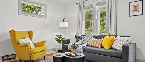 A living room with a yellow armchair, grey sofa, two black side tables, a floor lamp, and a patterned rug.