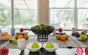 Una mesa con varias frutas y un bol de bolas verdes, ubicada en una habitación con ventanas grandes y vista a una casa.