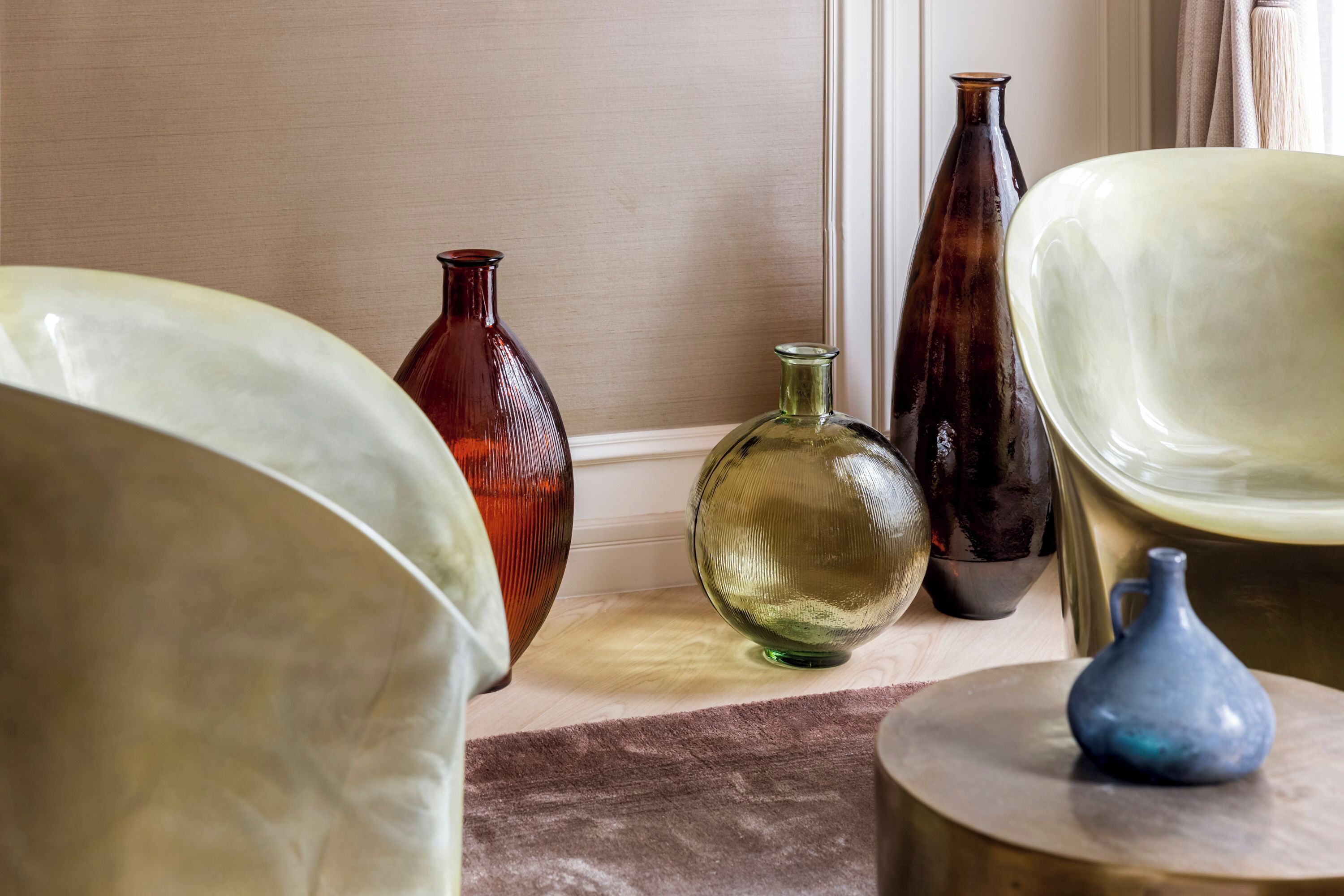 A room with a beige wall, a wooden floor, and a brown rug. There are several vases of different shapes and sizes on a wooden table.