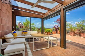An outdoor patio with a wooden ceiling, a white sofa with cushions, a wooden table, and potted plants.