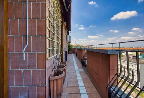 A narrow balcony with a tiled floor, potted plants, and a view of buildings in the distance.