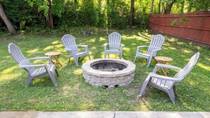 Un hoyo para fuego rodeado por cinco sillas Adirondack en un patio trasero con césped y una valla de madera al fondo.