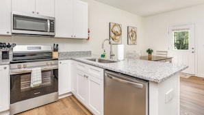 A modern kitchen with white cabinetry, a stainless steel dishwasher, a built-in oven, and a microwave. There is a granite countertop, a sink with a faucet, and a window providing natural light.