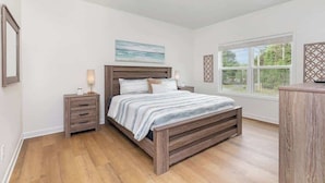 A bedroom with a wooden bed, a nightstand, a dresser, and a window with a view of greenery.