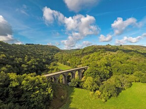 Uma ponte de pedra sobre um vale com vegetação exuberante e colinas ao fundo.