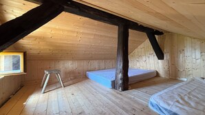 Pieni ullakkohuone, jossa on puuseinät ja -lattia, ikkuna, patja ja tikkaat.