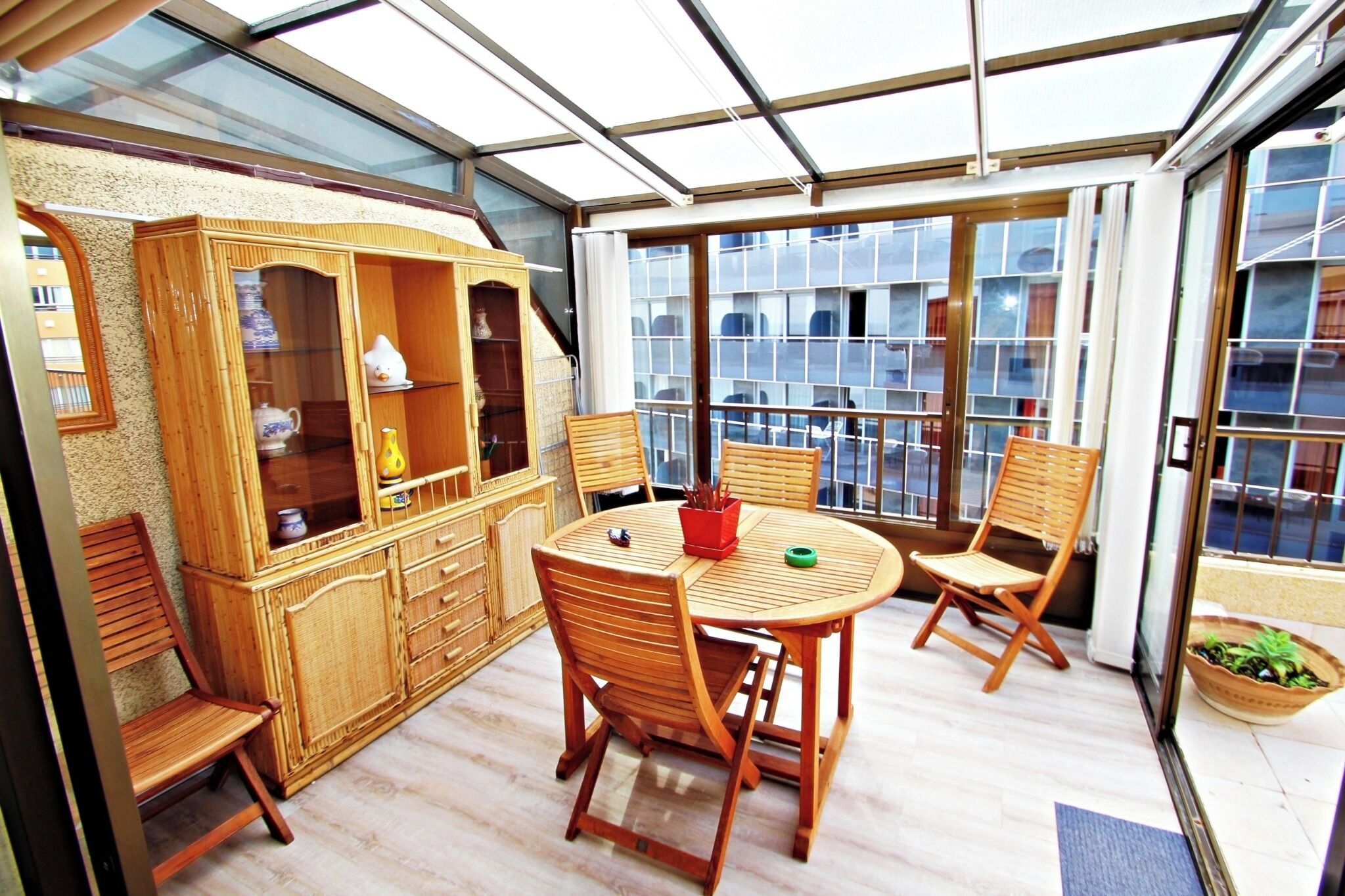 A sunlit room with a glass ceiling, wooden furniture, and a view of buildings through large windows.