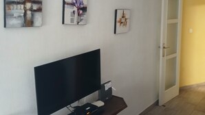 A room with a flat-screen TV on a wooden stand, a small table with a floral tablecloth, and three wall-mounted paintings. - Charming 2-bedroom apartment with WiFi, AC in fabulous Torrevieja (Torrevieja)