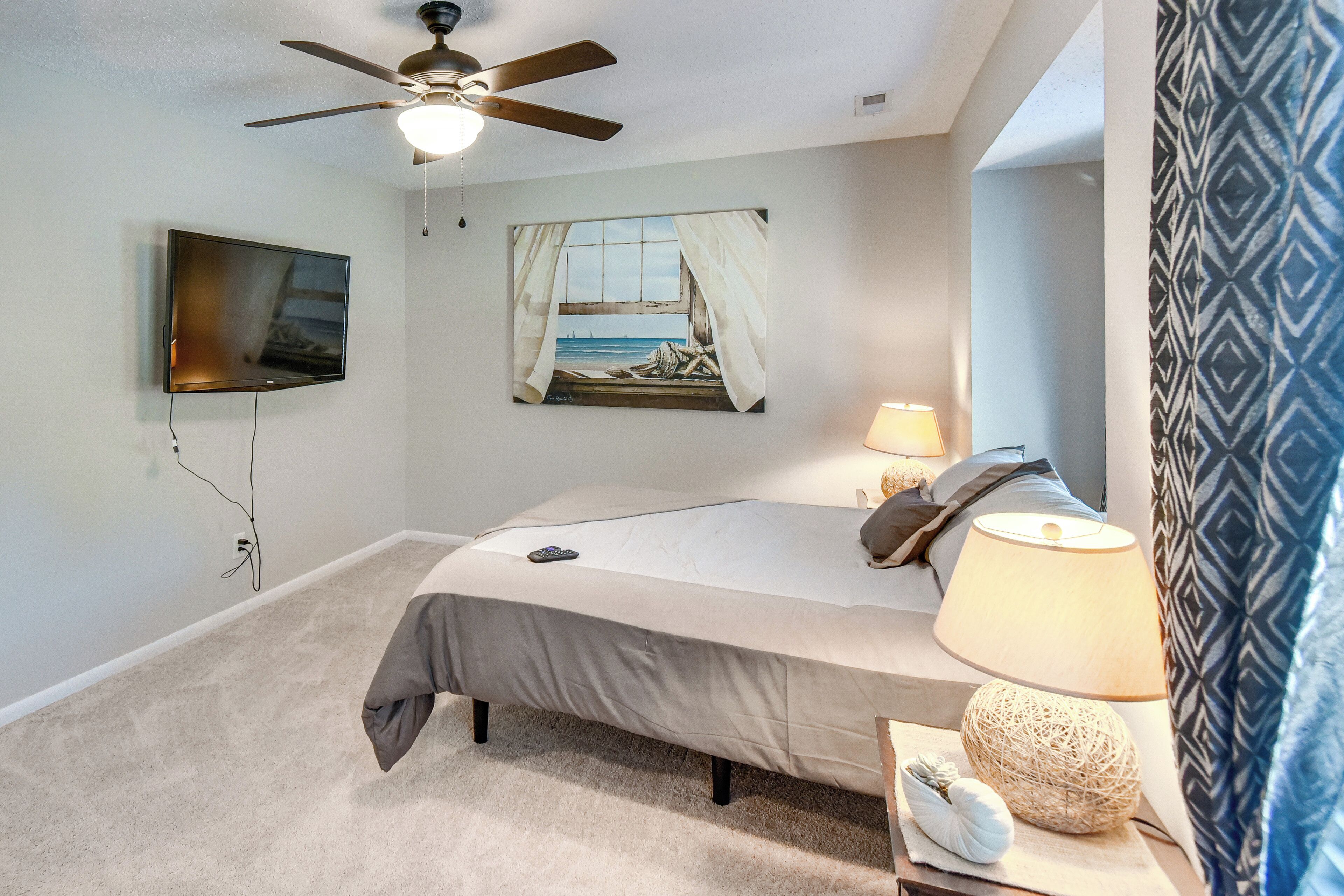 Une chambre à coucher avec un lit, un ventilateur de plafond, une télévision, un tableau, une lampe et un rideau à motifs.