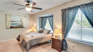 Ein Schlafzimmer mit Bett, Nachttischlampen, Deckenventilator, Fenster mit gemusterten Vorhängen und Wandbild.