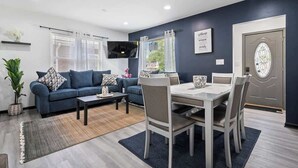 A modern living room with a blue sofa, a dining table with chairs, a TV mounted on the wall, and a door with a decorative glass panel.