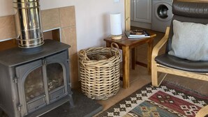 A cozy living room with a wood-burning stove, a wicker basket, a patterned rug, a comfortable chair, and a kitchen visible through an open doorway. - The Bothy -  a cottage that sleeps 4 guests  in 2 bedrooms (Lumphanan)