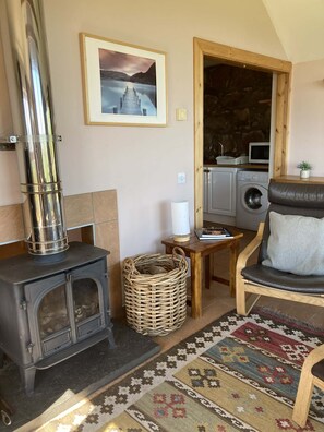 Una sala de estar acogedora con una estufa de leña, una cesta de mimbre, un tapete estampado, un sillón cómodo y una cocina visible a través de un arco.