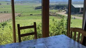 A table with a floral tablecloth and a book, two wooden chairs, and a view of a green landscape through large windows. - The Bothy -  a cottage that sleeps 4 guests  in 2 bedrooms (Lumphanan)