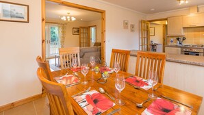 A dining area with a wooden table set for six, a kitchen in the background, and a door leading to a balcony. - Broombank Cottage - sleeps 6 guests  in 3 bedrooms (Inverness)