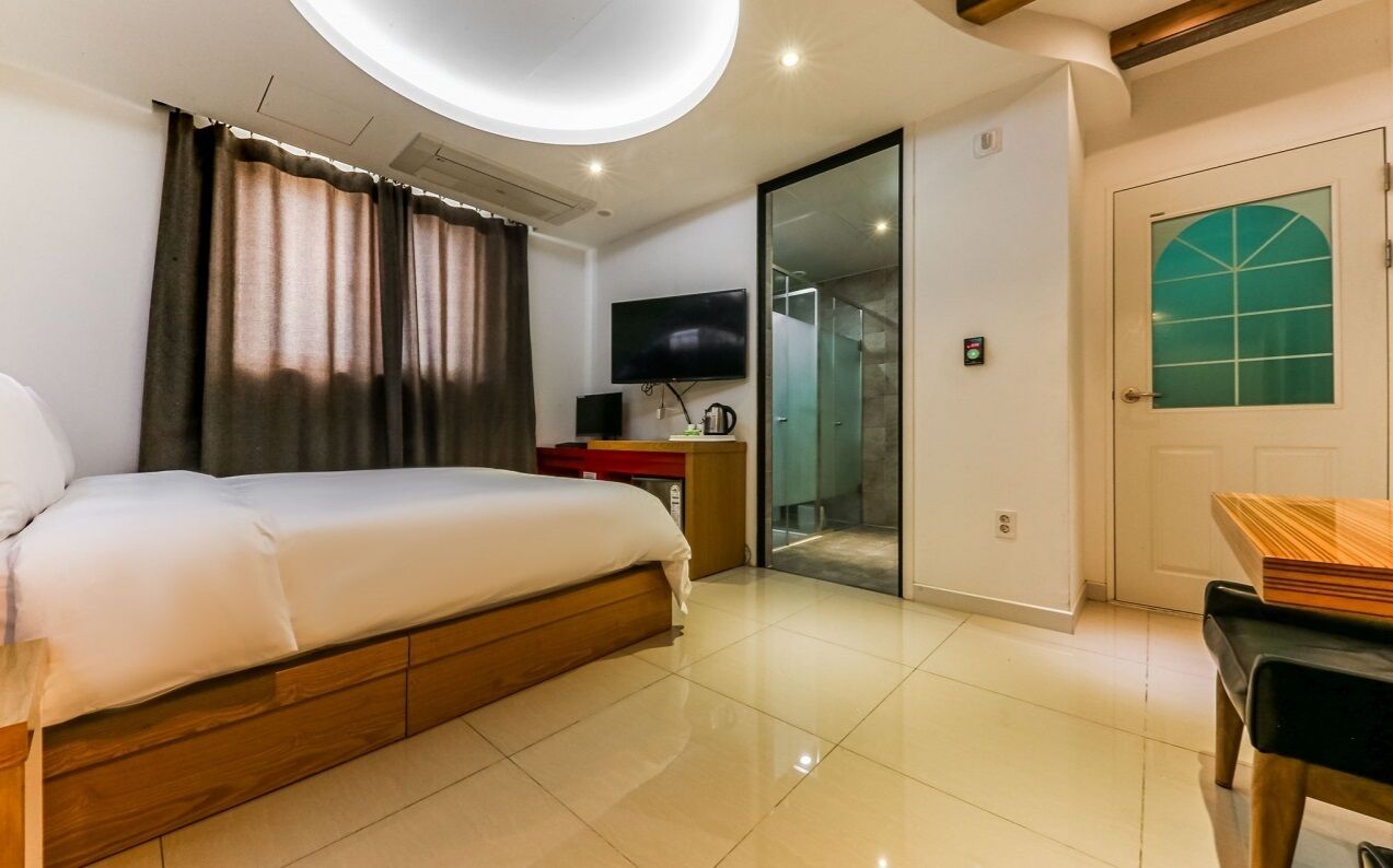 A modern hotel room with a large bed, a flat-screen TV, a desk with a chair, and a sliding glass door leading to a bathroom.