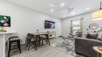 Una sala moderna con un sofá gris, una mesa de comedor con sillas, un televisor montado en la pared y un ventilador de techo.