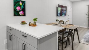 A modern kitchen with a white countertop, a dining area with a wooden table and chairs, and a TV mounted on the wall.